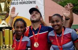 Duas garotas e um homem, todos com medalhas de ouro no peito, comemoram ao lado de troféu dourado.