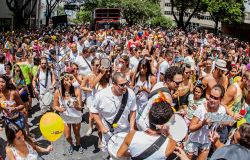Membros de um bloco de carnaval toca instrumentosmusicais; um pouco atrás, mais de trezentas pessoas acompanham; mais ao fundo há a presença de um caminhão de som.