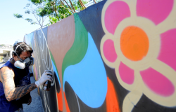 Homem com máscara de segurança e luva grafita muro com flores e cores vibrantes, durante o dia.