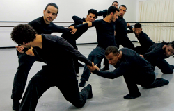 Oito homens vestidos de preto interagem em apresentação de dança.