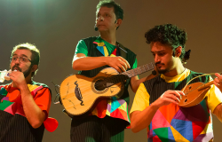 Três homens se apresentam com camisetas coloridas e instrumentos musicais.