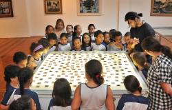 Mais e trinta crianças observam peça de museu, acompanhados por mediador.