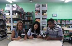 três estudantes lêem livros, ao fundo estante de biblioteca com vários livros. 