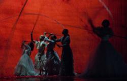 Em cena teatral, quatro mulheres de vestido longo movimentam astes, suas sobras são projetadas no fundo vermelho. 