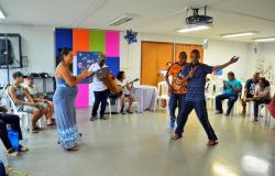 Mais de dez pessoas fazem parte de sarau com dança e violão em sala com cadeiras. 