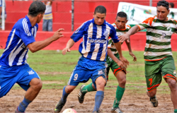 Quatro jogadores de futebol, dois de uniforme azul e dois de uniforme verde, disputam uma bola em uma partida da copa Centenário de Futebol Amador.