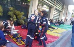 TRês membros da equipe do SAMU atendem a mais de dez pessoas deitadas no chão da calçada, simulando passar mal devido a incêndio simulado. 