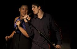 Dois jovens, um homem e uma mulher, cantam com microfones, em cena teatral de fundo escuro.