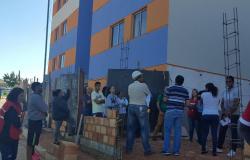 Cerca de dez pessoas, uma delas com capacete, prédio azul e laranja, cuja construção foi recém concluída, durante o dia.