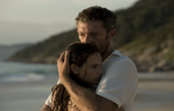 Homem abraça garota com praia ao fundo, durante o dia. Imagem do filme "À Deriva", em cartaz em mostra do MIS Cine Santa Teresa. 