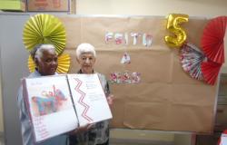 Duas idosas usuárias do centro de saúde seguram o livro artesanal a frente de um cartaz com os dizeres "Feito a mão"