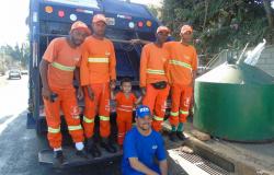 Criança ao lado de cinco profissionais da Limpeza Urbana na traseira do caminhão de lixo da SLU