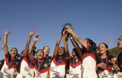 Equipe de futebol feminino sub-17 doTupinambás, todas as jogadoras com univorme e medalha, erguem a taça da Copa Centenário.