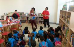 Cerca de vinte crianças sentadas no chão enquanto três adultos contam e interpretam histórias em uma biblioteca.