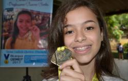 Garota segura medalha da OBMEP; ao fundo, Banner da Obmep com foto da mesma garota.