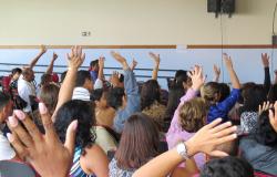 A imagem mostra cerca de 30 pessoas sentadas em um auditório, com as mão levantadas, em uma votação.