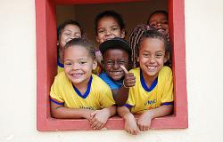 A foto mostra seis crianças, de quatro e cinco anos, em uma janela, sorrindo para a câmera.