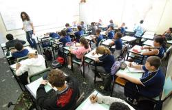 Foto mostra sala de aula com alunos com idade aproximada de 11 anos, sentados em carteiras. Na frente da sala há uma professora em pé, próxima ao quadro branco.