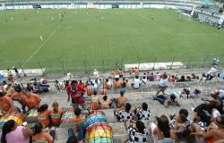 A foto mostra a vista de um campo de futebol a partir da arquibancada, onde é possível ver cerca de cinquenta pessoas