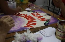 Foto de uma oficina de artesanato. Há mãos de mulheres bordando e o texto pintado em um tecido: CEAM Benvinda