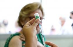 Imagem de seringa e agulha com frasco de vacina contra a gripe. Ao fundo, idosa aguardando a aplicação